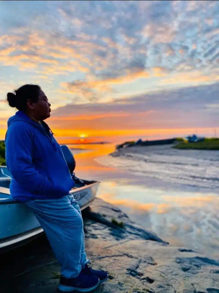 A woman leans against a small boat against a sunset ocean backdrop.